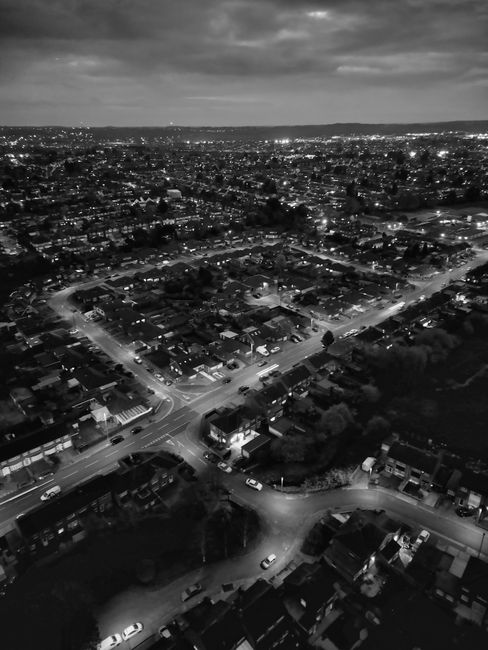 Black and white aerial view of a residential neighbourhood at night with numerous houses and streets illuminated by streetlights, showing a mix of detached and semi-detached homes with roof tiles and small gardens. The scene captures the orderly layout of the area, with visible roads and parked cars, under a cloudy sky. The image emphasizes city lighting and the quiet, clean appearance of the area, suitable for highlighting the importance of surface cleaning and maintenance in domestic environments. Mentioned in the context of [COMPANY_NAME]’s professional cleaning and sanitisation services near Cassiobury Park Watford, this image showcases the importance of thorough cleaning for a well-maintained residential community.