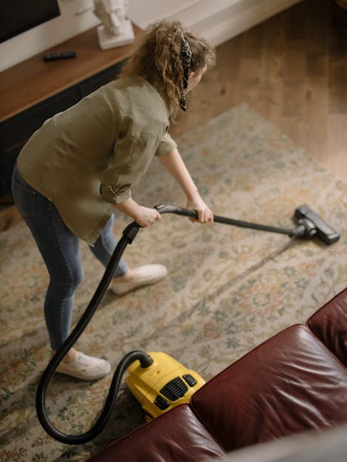 A woman with curly hair, dressed in a beige jacket, blue jeans, and white shoes, is performing surface cleaning on a patterned area rug using a yellow vacuum cleaner with a black hose attachment. She is in a living room with warm wooden flooring and a wooden sideboard against the wall, which holds a small decorative item. The room is well-lit with natural light, highlighting the cleanliness and condition of the rug, which appears freshly vacuumed and free of dust. The setting emphasizes domestic cleaning and maintenance services provided by Watford Carpet Cleaning, with a focus on deep cleaning and sanitisation of carpets and rugs in residential spaces near Cassiobury Park Watford.