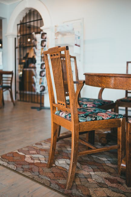 A wooden dining chair with a floral patterned upholstered seat positioned next to a matching wooden table on a decorative rug in a well-lit room. In the background, there is an open archway with a wrought iron gate, leading to another area of the space. The room features light-colored walls and wooden flooring. Visible in the background is a stand with various brochures or notices and additional wooden chairs, indicating a domestic or hospitality setting. The scene emphasizes the clean condition of the furniture and the overall tidy appearance of the space, reflecting professional domestic surface cleaning, deep cleaning, and sanitisation services provided by Watford Carpet Cleaning as featured in their success story for Vicarage Road carpet and upholstery cleaning.